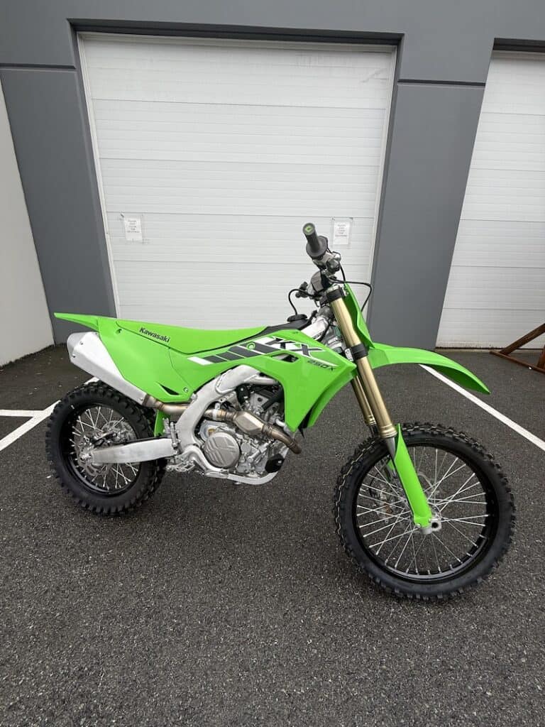 Green Kawasaki KX250X dirt bike parked outside with garage doors in background.